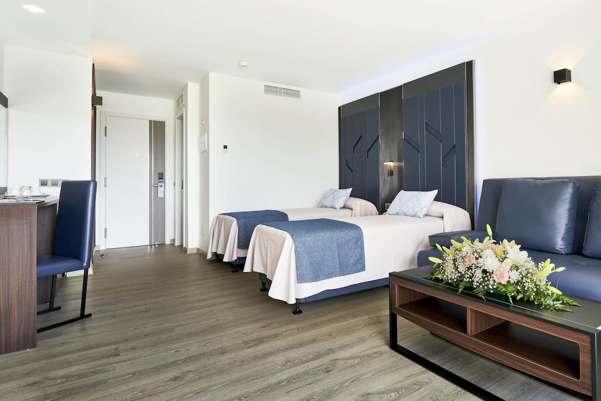 Une chambre d’hôtel moderne avec un plancher en bois, un lit doté d’une tête de lit bleue, un canapé bleu, une table basse en bois ornée de fleurs et un bureau avec une chaise.