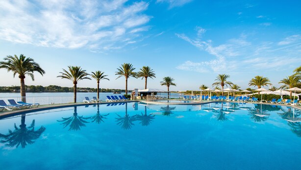A swimming pool with blue water, surrounded by palm trees and lounge chairs. - Hotel BelleVue Club (Alcúdia)