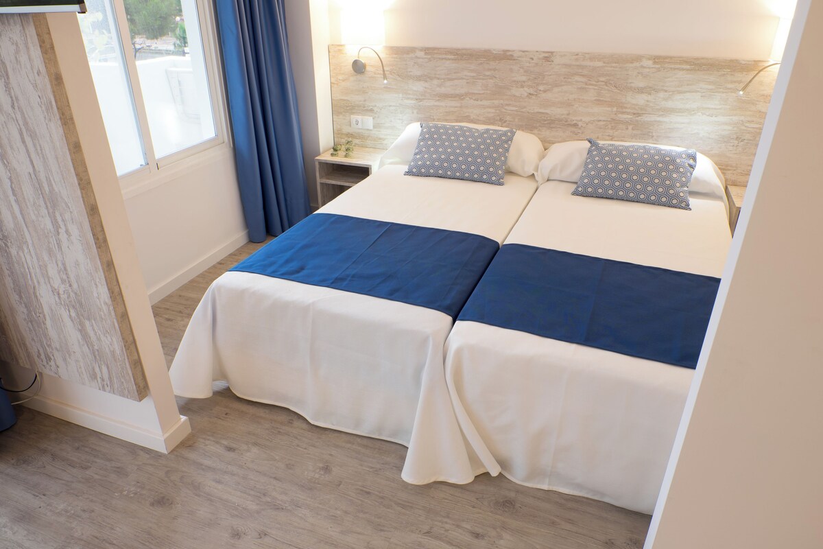 A double bed with a blue and white bedspread, a bedside table with a lamp, and a window with curtains.