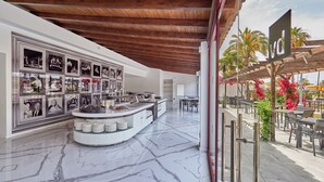 A modern hotel lobby with a marble floor, a central buffet area, and a seating area with outdoor views. - Hotel BelleVue Club (Alcúdia)