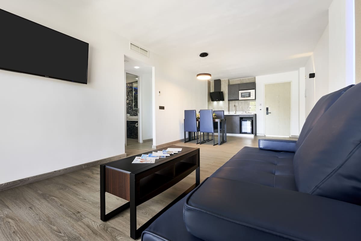 Un salon moderne avec un canapé en cuir noir, une table basse sur laquelle sont posés des papiers, un téléviseur à écran plat et un coin repas avec des chaises bleues, le tout donnant sur une cuisine en arrière-plan.