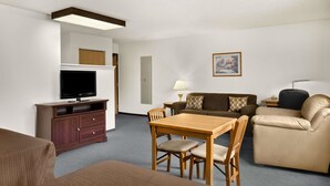 Habitación de hotel con un mueble bajo de televisión de madera, un televisor de pantalla plana, una mesa de comedor de madera con cuatro sillas, un sofá marrón, un sillón de cuero color beige y una mesita de noche con una lámpara.