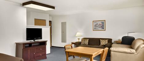 Une chambre d’hôtel comprenant un meuble TV en bois, un téléviseur à écran plat, une table à manger en bois avec quatre chaises, un canapé marron, un fauteuil en cuir beige et une petite table d’appoint avec une lampe.