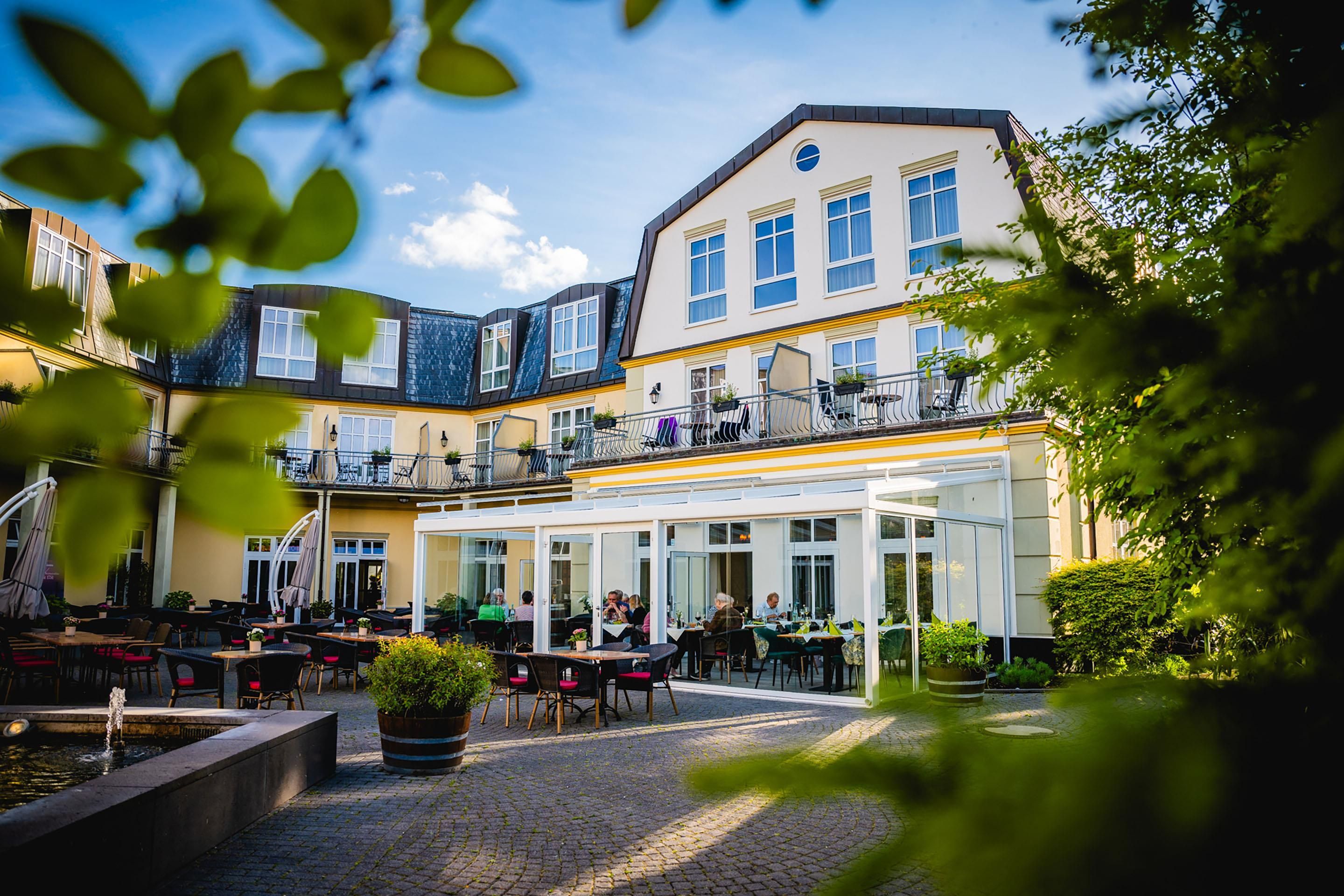 An outdoor café area with tables and chairs, a water feature, and a building with multiple windows and balconies.