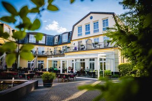 Een terras met tafels en stoelen, een waterpartij, en een gebouw met meerdere ramen en balkons.