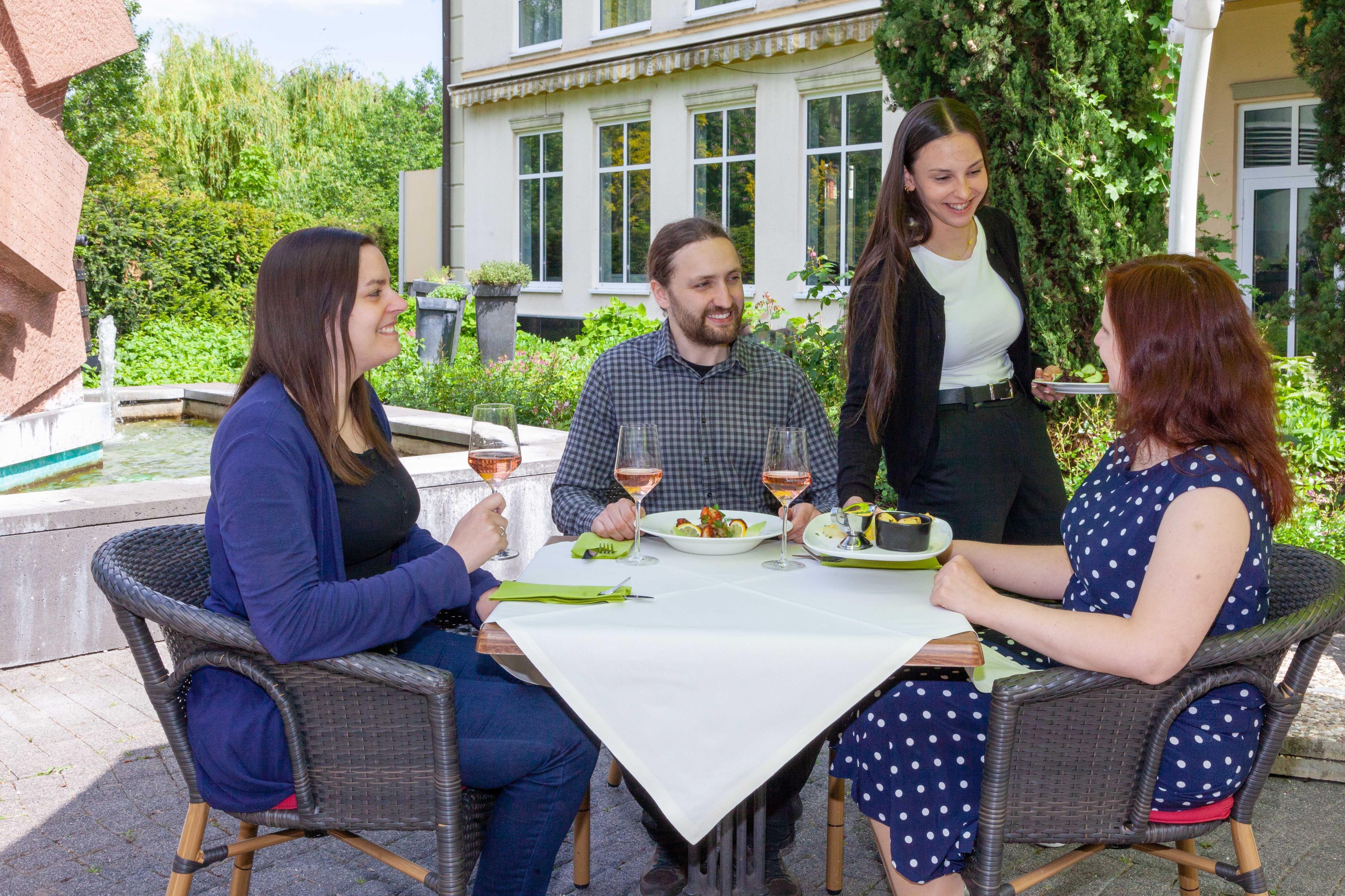 Un grupo de personas cenando al aire libre en una mesa con mantel blanco, rodeado de follaje y un edificio al fondo.