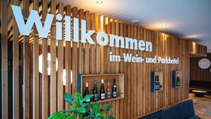 Un espace d’accueil avec un mur en bois et une enseigne indiquant : « Bienvenue au Wein- und Parkhotel ». On y trouve plusieurs bouteilles de vin exposées sur une étagère, une plante en pot et des coins salon.