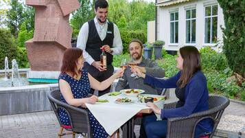 Eine Gruppe von Personen speist im Freien an einem Tisch, während ein Kellner Getränke serviert.