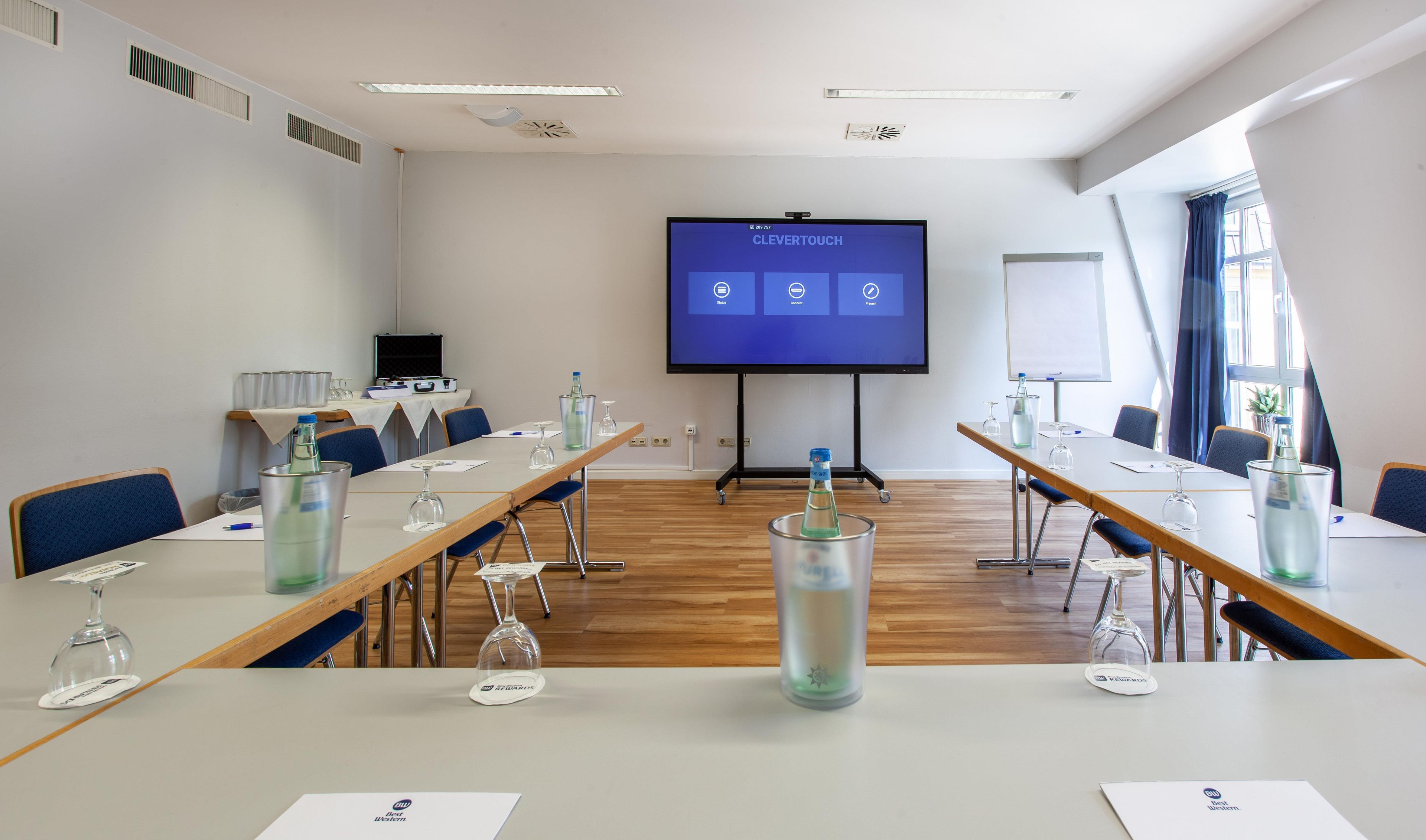 A conference room with a large screen displaying a presentation, wooden tables with chairs, and a whiteboard.
