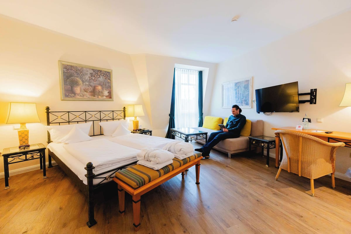 Ein Hotelzimmer mit einem Bett, zwei Nachttischen, einem Schreibtisch, einem Sessel, einem Fernseher und einem Fenster mit Vorhängen.