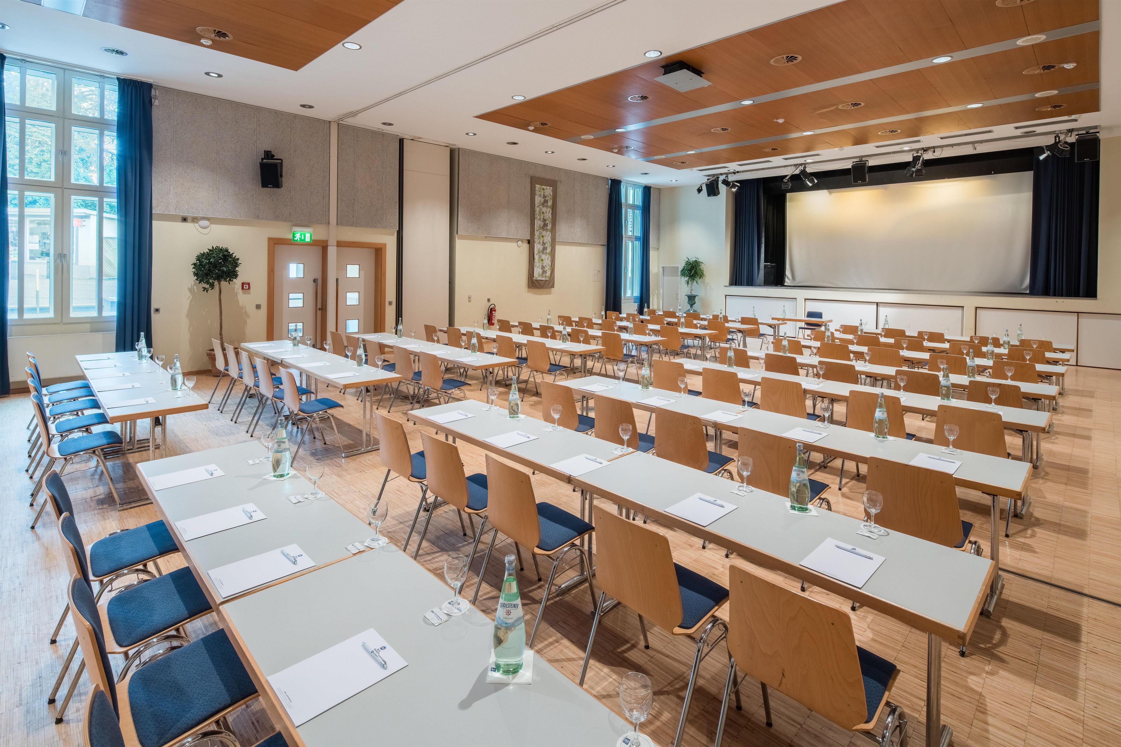 Ein Konferenzraum mit Reihen von Tischen und Stühlen, einer Bühne mit Bildschirm und einem großen Fenster mit Vorhängen.