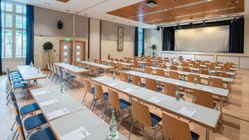 Ein Konferenzraum mit Reihen von Tischen und Stühlen, einer Bühne mit Bildschirm und einem großen Fenster mit Vorhängen.