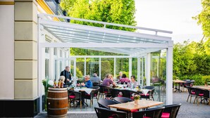 Een terras buiten met tafels, stoelen, een glazen overkapping en een vat.