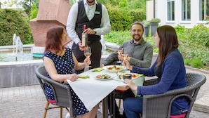 Un groupe de personnes est assis à une table à l’extérieur, où elles prennent leur repas, et un serveur leur apporte des boissons.