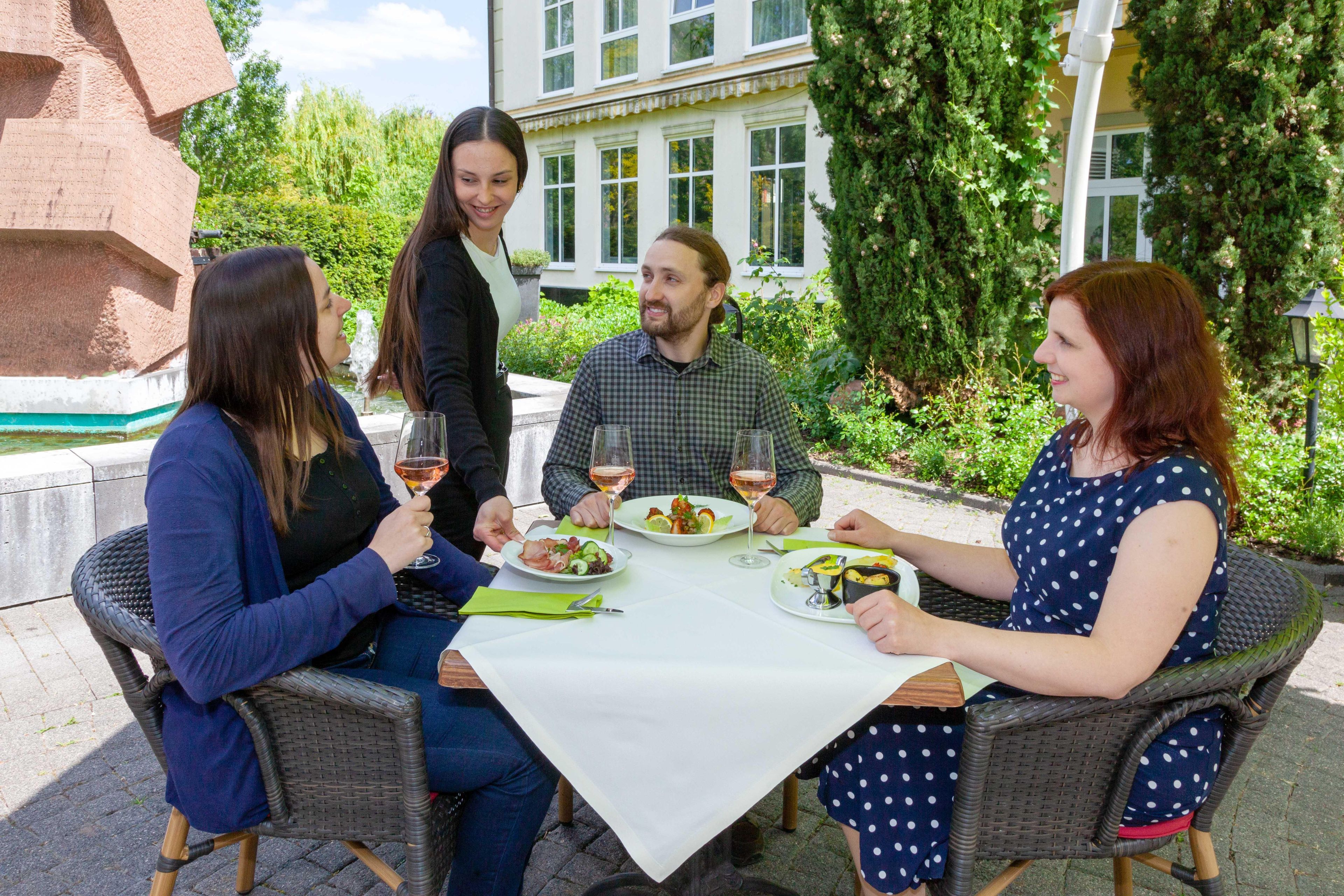 Un grupo de personas cenando al aire libre en una mesa con vista a un edificio y follaje.