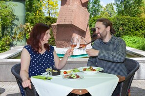 Twee personen dineren aan een buitentafel met uitzicht op een groot stenen gebouw en groen.