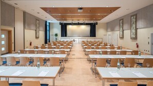 Une salle de conférence avec des rangées de tables, des chaises et une scène équipée d’un écran.
