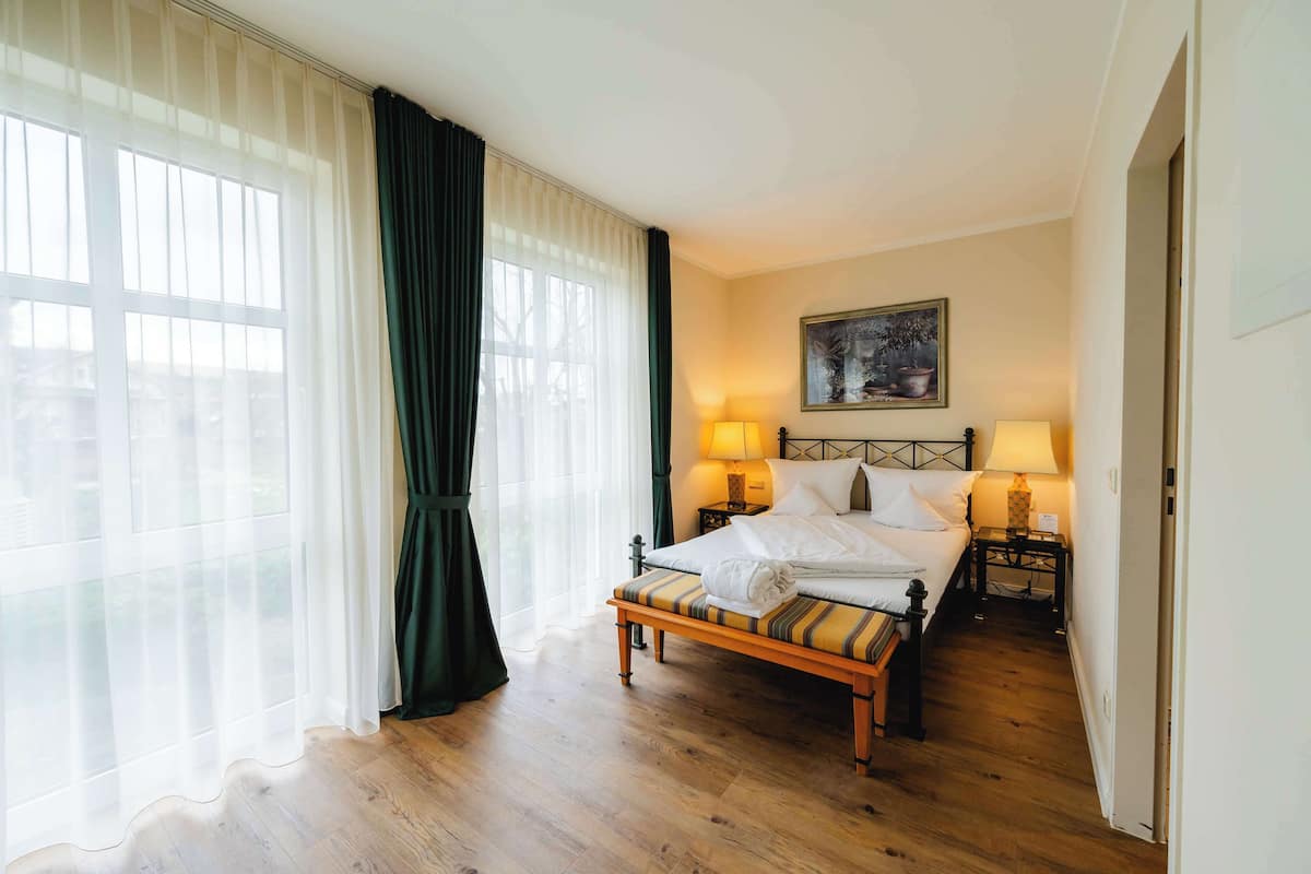 Ein ordentlich eingerichtetes Schlafzimmer mit Bett, Nachttischlampen, einer Bank und einem großen Fenster mit durchsichtigen Vorhängen.