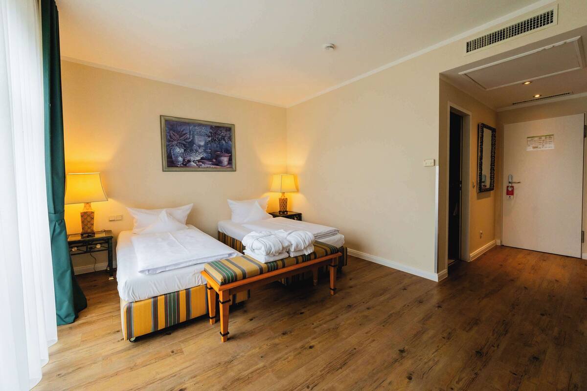 Ein Hotelzimmer mit zwei Betten, einer Sitzbank, einem Gemälde und einer Tür zu einem anderen Raum.
