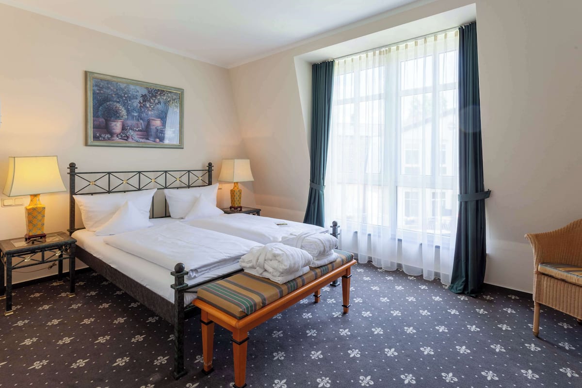 Ein Hotelzimmer mit Doppelbett, zwei Nachttischen mit Lampen, einer Bank, einem Sessel und einem Fenster mit Vorhängen.