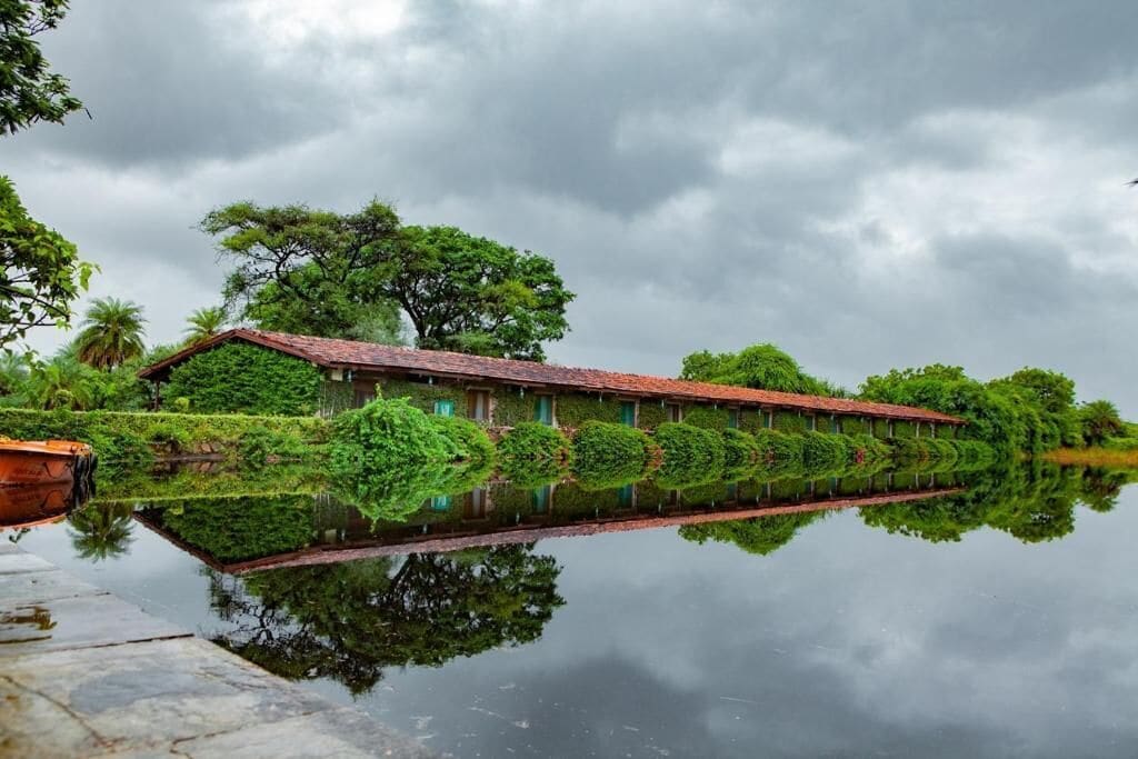 A building with a red roof and green foliage, reflecting in a calm body of water.