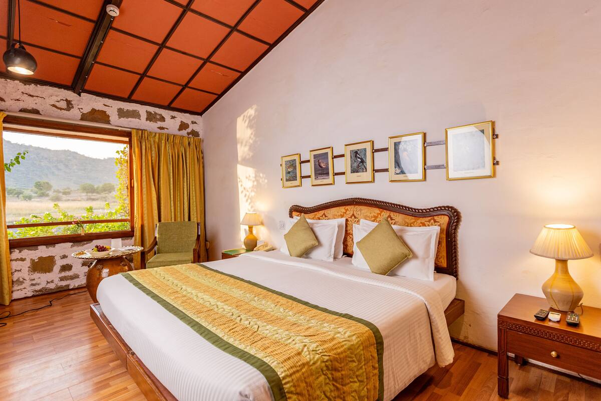 Habitación de hotel con una cama grande, vistas al exterior, cabecera de madera, mesita de noche con lámpara y cuadros en la pared.
