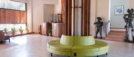 Un hall amplio con una zona de estar circular central, un mostrador de recepción de madera, plantas en macetas y una escalera con una estatua.