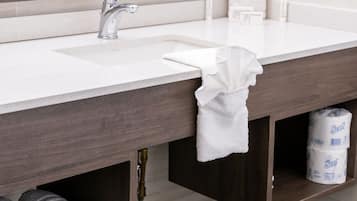 Casa de banho com lavatório branco, espelho e suporte para toalhas. Toalhas dobradas e papel higiénico estão nas prateleiras.