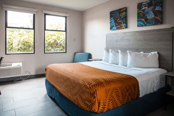 A modern hotel room with a large bed, two windows with blinds, a chair, and artwork on the wall.