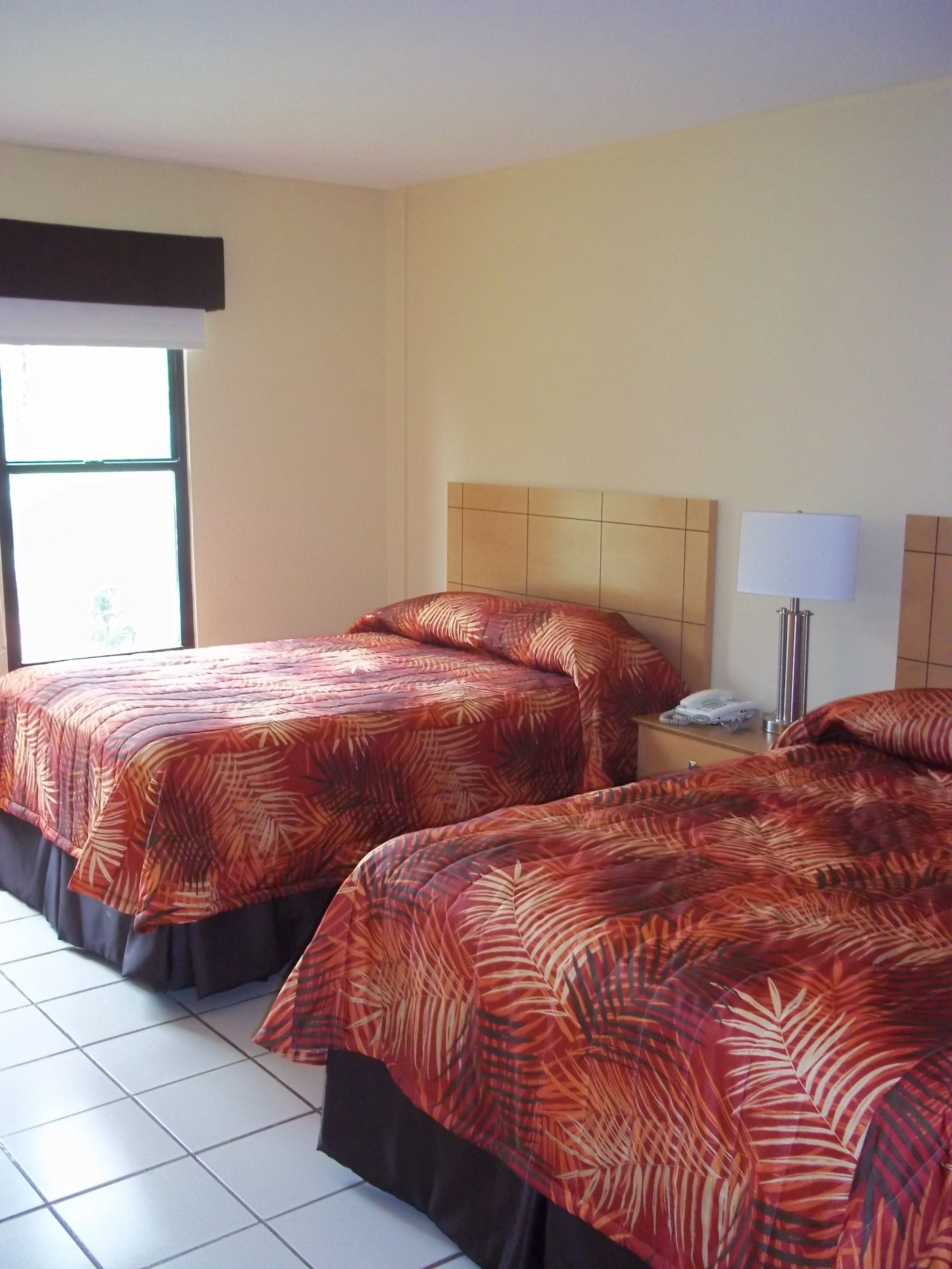 A hotel room with two beds, each with a patterned bedspread, a bedside table with a lamp, and a window with blinds.