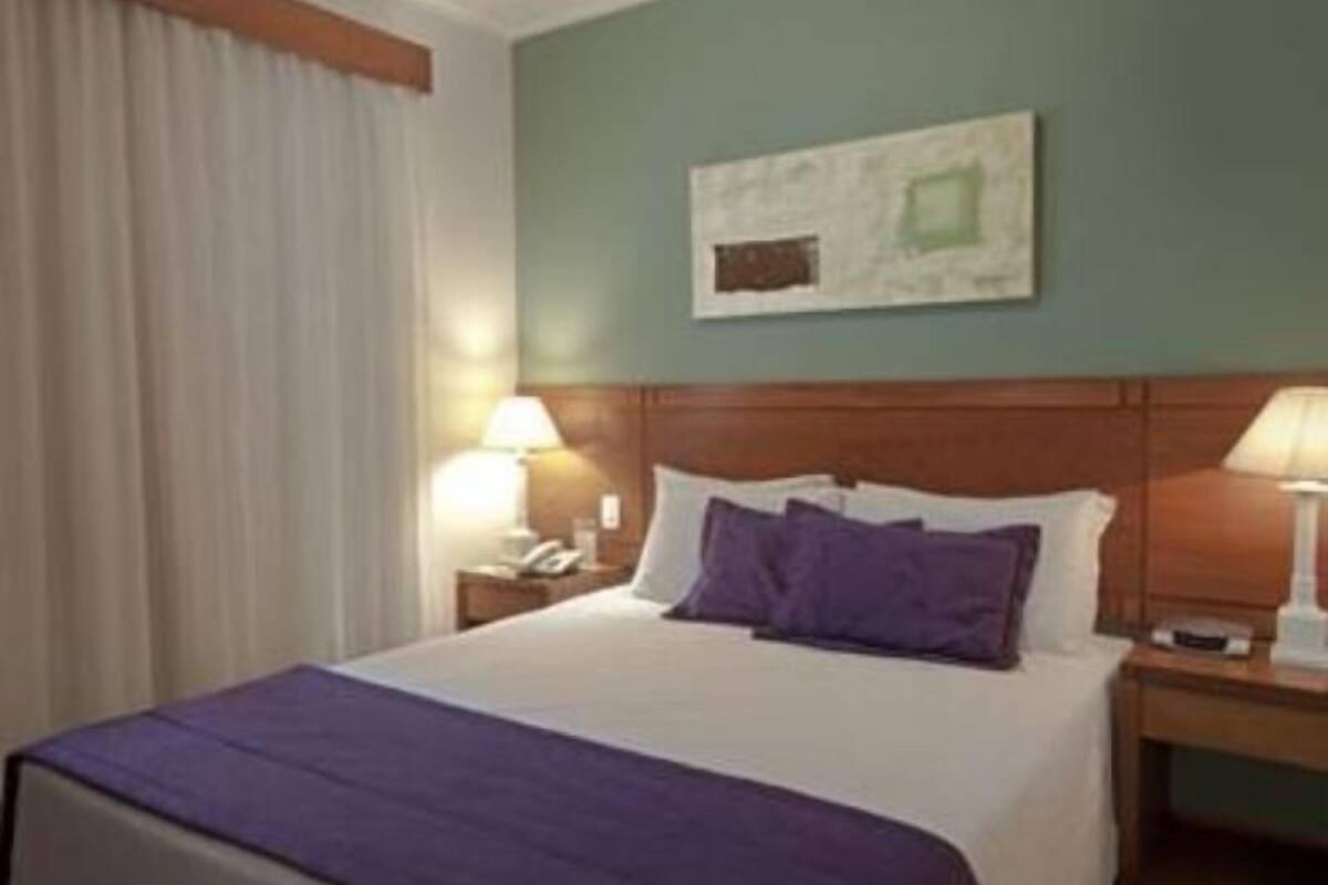 Habitación de hotel con cama, dos mesitas de noche, lámpara, un cuadro en la pared y ventana con cortinas.