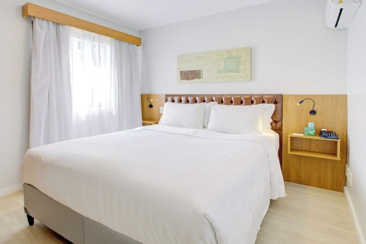 A hotel room with a large bed, a wooden headboard, a bedside table with a lamp, a nightstand with a phone, and a painting on the wall.
