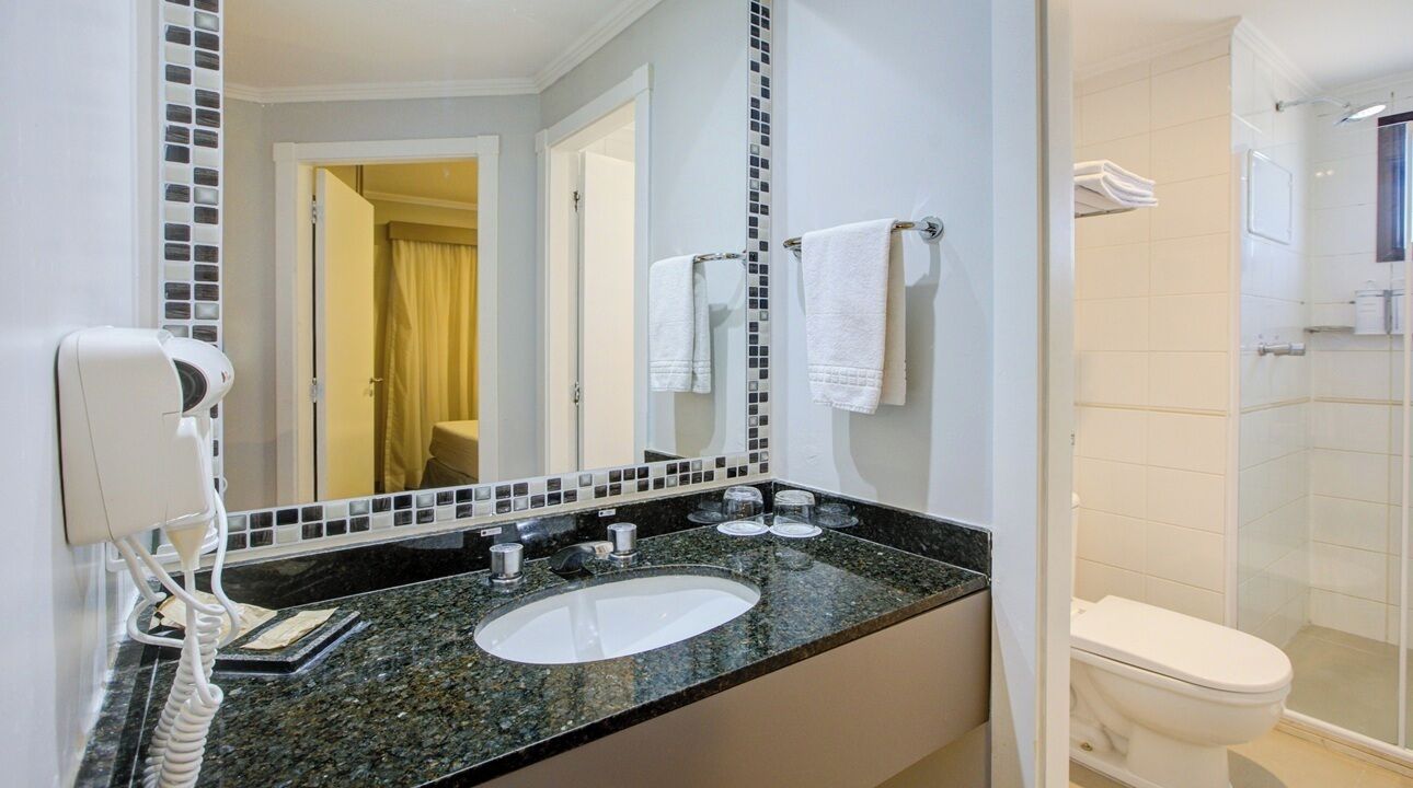 A bathroom with a granite countertop, a white sink, a mirror with a mosaic backsplash, a white toilet, and a glass shower enclosure.