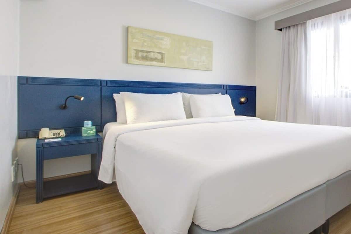 A hotel room with a large bed, a bedside table with a lamp, a nightstand, and a window with curtains.
