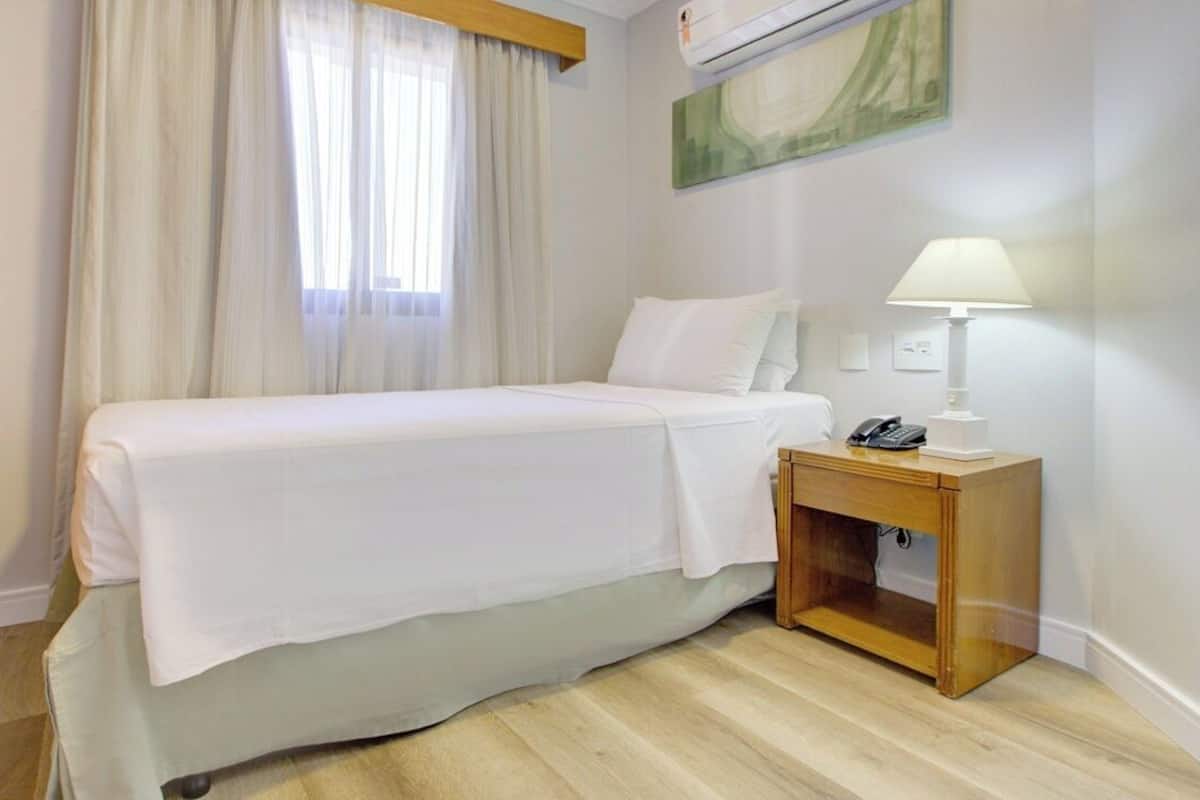 A single bed with white bedding, a bedside table with a lamp, and a wall-mounted air conditioner.