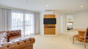 Une chambre d’hôtel moderne avec un canapé en cuir, une table à manger avec des chaises, un téléviseur à écran plat fixé sur un panneau en bois, et un lit en arrière-plan.