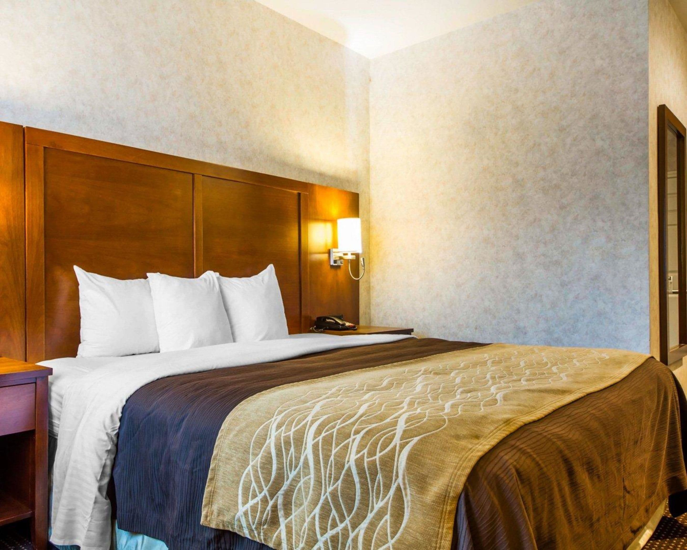 A hotel room with a wooden headboard, a bed with white and brown bedding, a nightstand with a phone, and a wall-mounted light fixture.