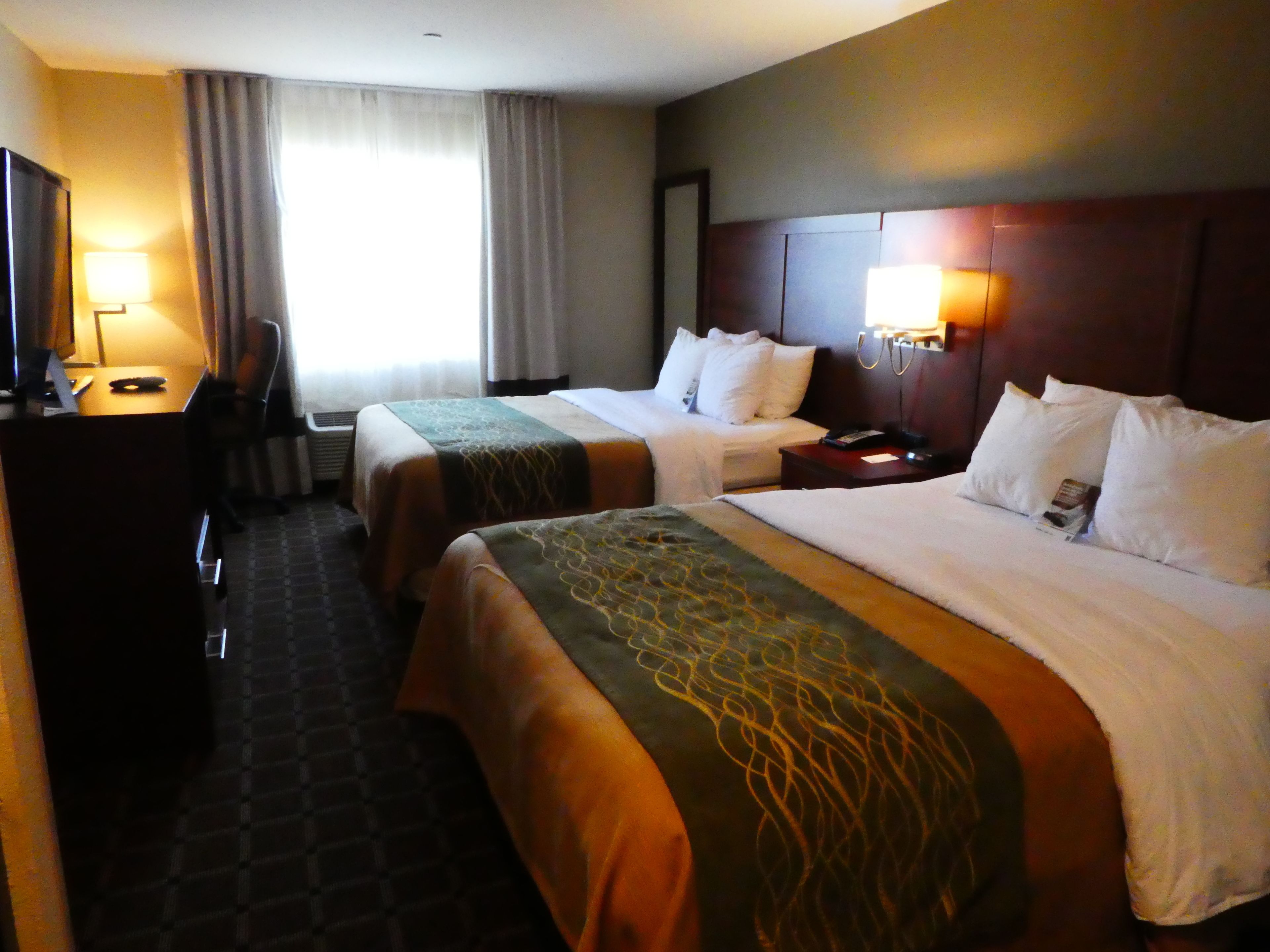 A hotel room with two beds, a desk, a chair, a lamp, and a window with curtains.