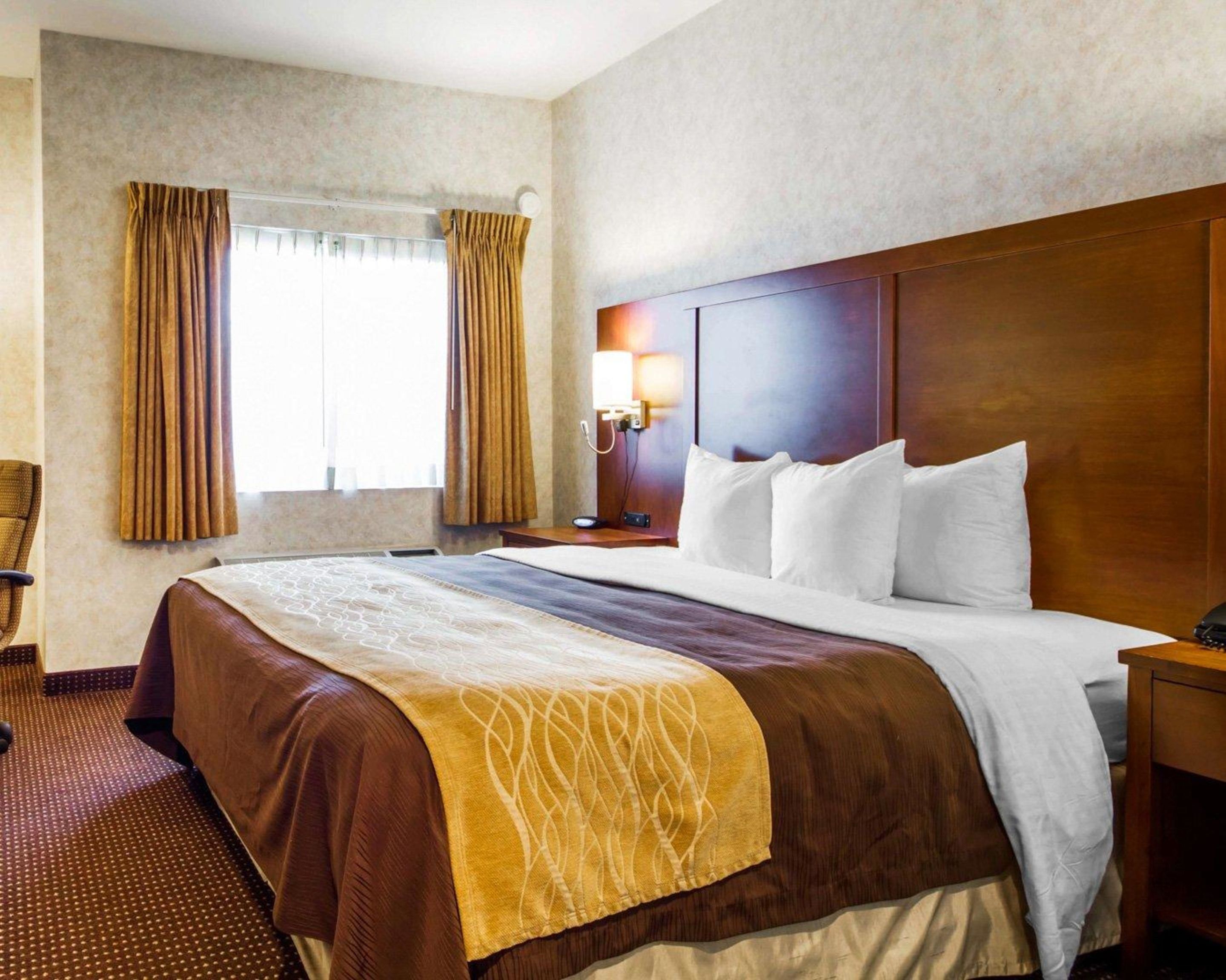 A hotel room with a large bed, a wooden headboard, a nightstand with a lamp, and a window with curtains.