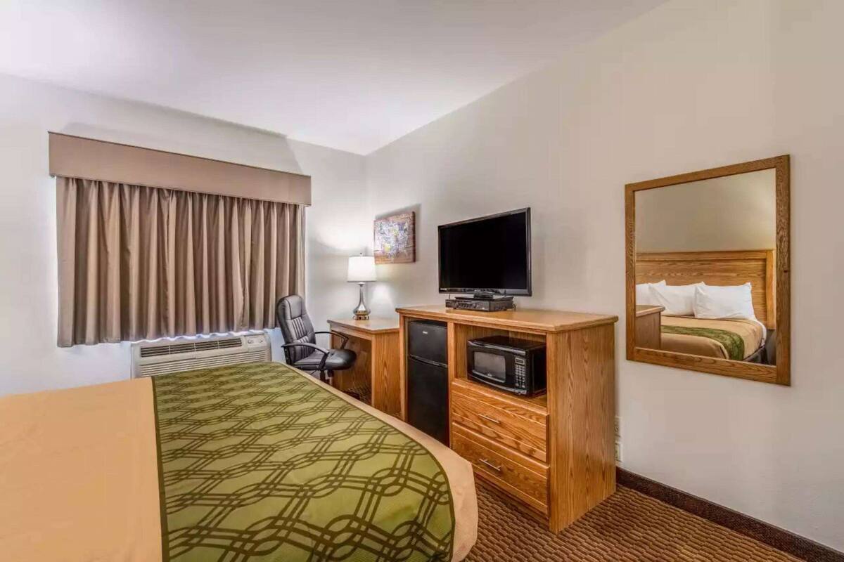 Une chambre d’hôtel comprenant un lit, un bureau avec un ordinateur, une télévision, un miroir et une fenêtre avec des rideaux.