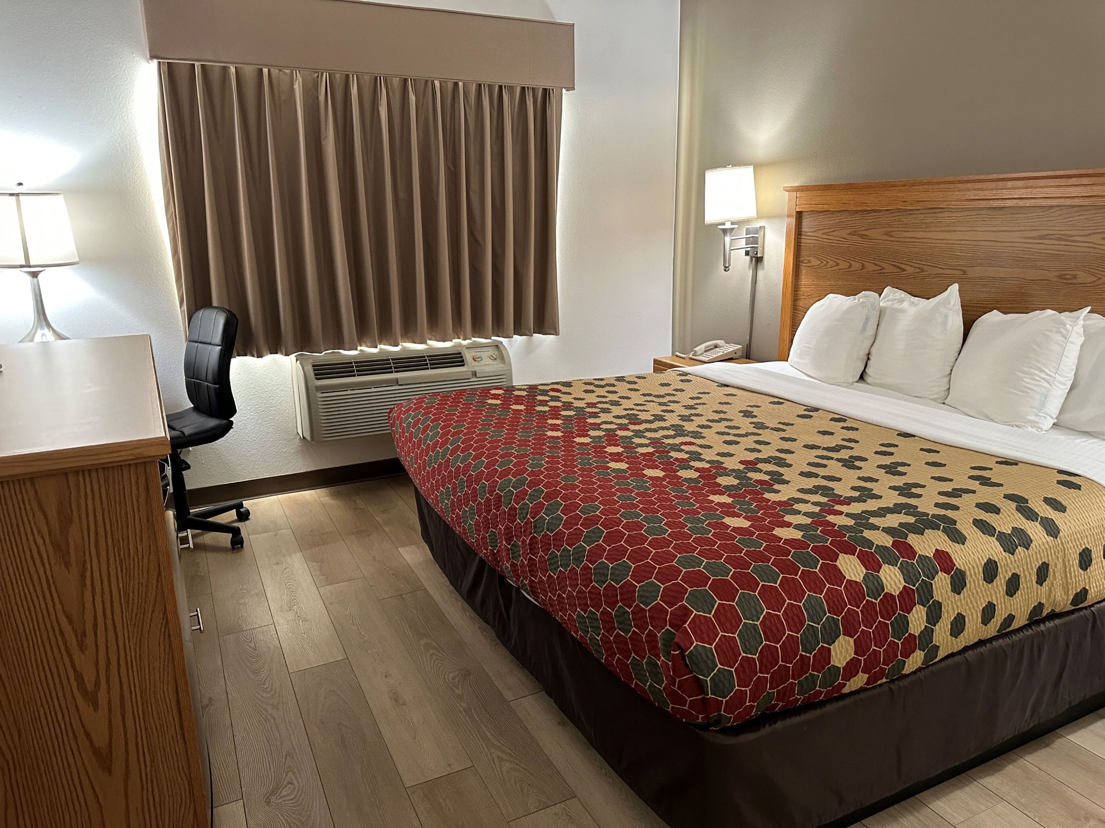 A hotel room with a bed, a desk, a chair, a lamp, a phone, and an air conditioning unit.