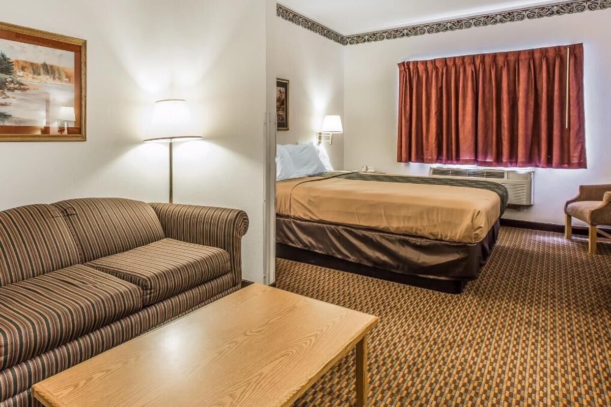 A hotel room with a sofa, a wooden table, a bed with a brown bedspread, a window with red curtains, and a painting on the wall.