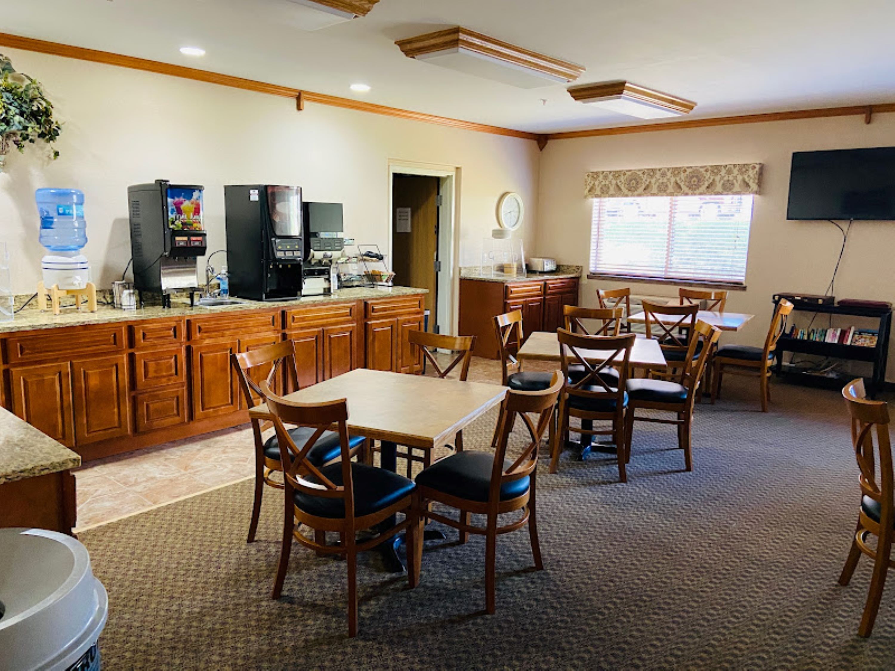 A spacious kitchen with wooden cabinets, a dining area with wooden chairs, a water dispenser, and a flat-screen TV mounted on the wall.