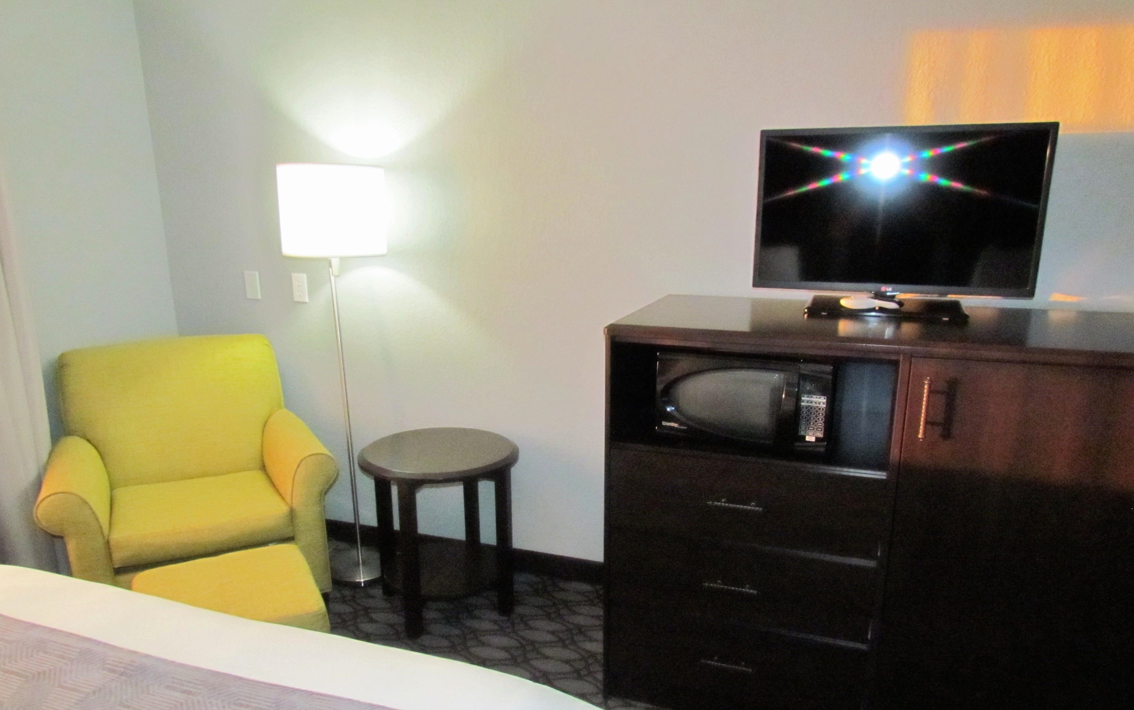Habitación de hotel con un sillón amarillo, una mesita redonda pequeña, un televisor sobre un mueble bajo y una lámpara de pie.