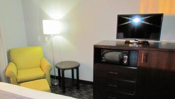 Habitación de hotel con un sillón amarillo, una mesita redonda pequeña, un televisor sobre un mueble bajo y una lámpara de pie.