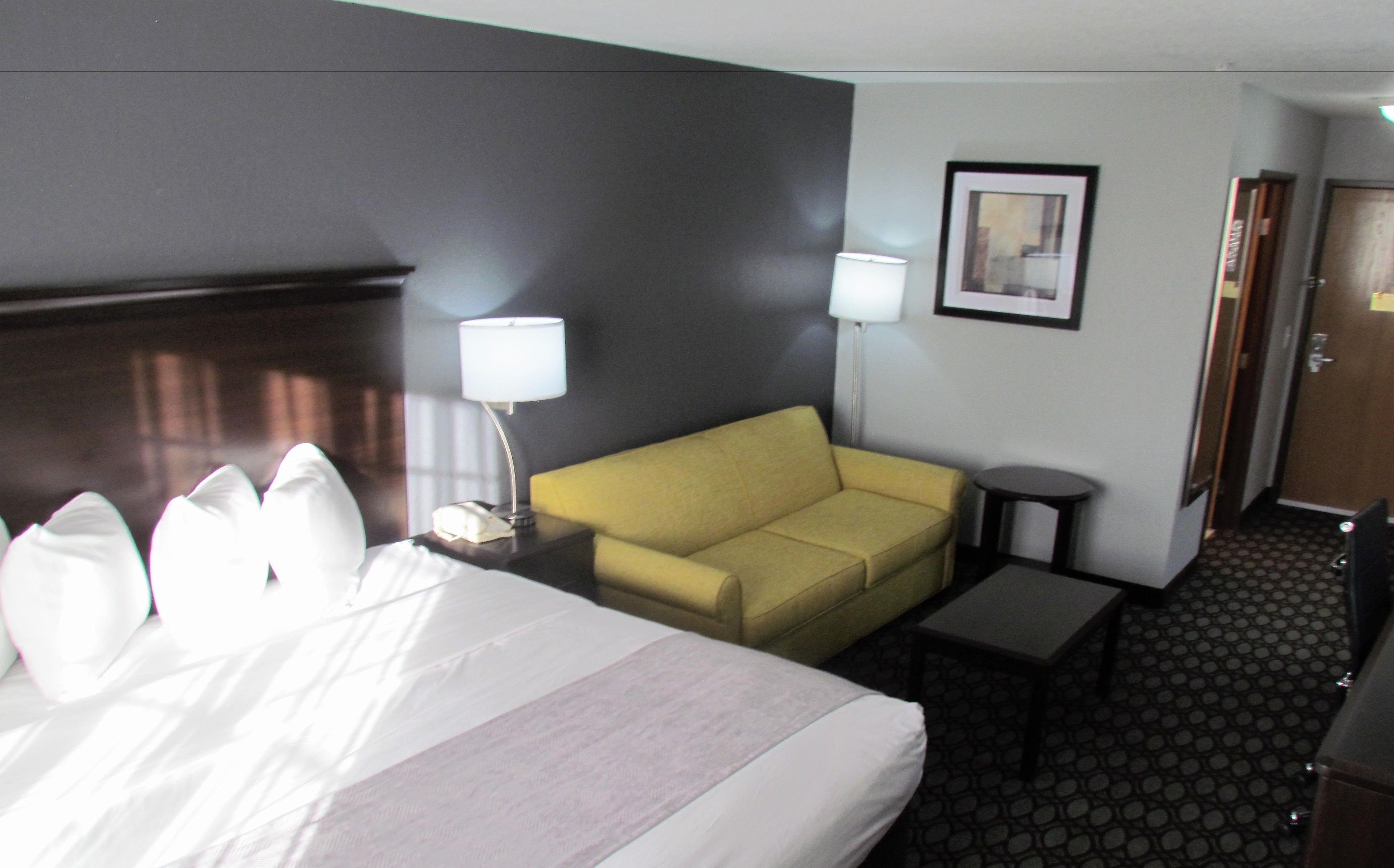 A hotel room with a bed, a yellow sofa, a small table, a lamp, and a framed picture on the wall.