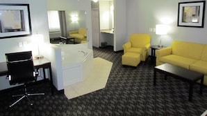 A hotel room with a bathtub, two yellow armchairs, a desk with a chair, a small table, and a lamp.