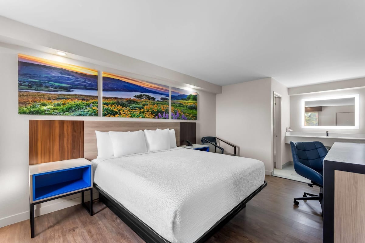 A modern hotel room with a large bed, a desk, and two paintings on the wall.