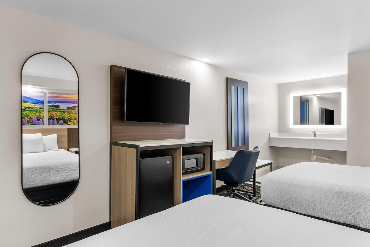 A hotel room with two beds, a desk with a flat-screen TV, a minibar, a mirror, and a window.