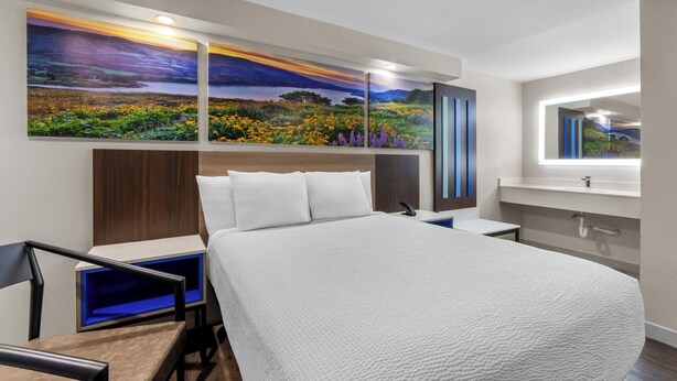 A modern hotel room with a large bed, a nightstand, a desk, and a large painting of a landscape.